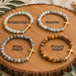 May include: Four beaded bracelets arranged in a square, each featuring round stone beads and a gold-toned cross charm. The bracelets are labeled Amazonite, Howlite, Atlas Jasper, and Picture Jasper, showcasing light blue, white marbled, grey marbled, and brown tones.