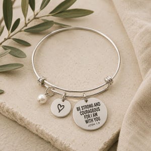 May include: A silver-toned expandable bangle bracelet featuring three charms: a small white pearl, a circular disc with an engraved heart symbol, and a larger circular disc engraved with the text BE STRONG AND COURAGEOUS FOR I AM WITH YOU JOSH 1:9.