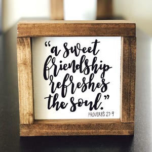 May include: A square wooden sign with a dark brown stained frame and a white center panel. The text in black script reads, "a sweet friendship refreshes the soul." and includes the citation "PROVERBS 27:9" in the bottom right corner.