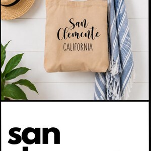 May include: A tan canvas tote bag featuring the black printed text San Clemente CALIFORNIA. The bag has two matching fabric handles and a rectangular shape. The design is centered on the front of the bag with a script font for the city name.