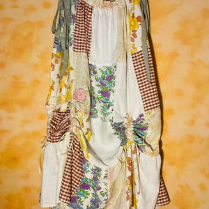 May include: A floor-length patchwork skirt featuring a variety of fabric patterns including brown and white gingham, yellow floral prints, and purple floral designs. The skirt has an elasticated waist with side ties and adjustable drawstring ruching.