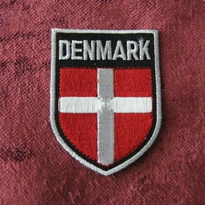 May include: An embroidered patch featuring the Danish flag, with the word "DENMARK" in white lettering on a black background. The patch is shaped like a shield.