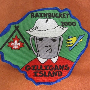 May include: Embroidered patch with a green and blue border. The patch features a cartoon bucket wearing a hat, with the text "RAINBUCKET 2000" above and "GILLIGANS ISLAND" below. A maple leaf and a stylized hand are also included.
