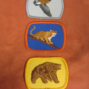 May include: Three embroidered patches featuring a goose in flight on a white background, a mountain lion on a blue background, and a brown bear on a yellow background.