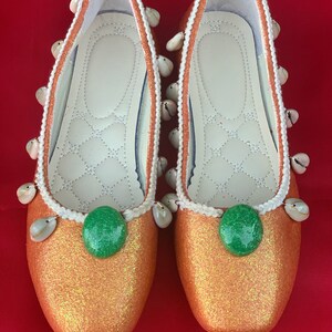 May include: A pair of orange glitter ballet flats with white trim and seashell embellishments. Each shoe has a green gem in the center.