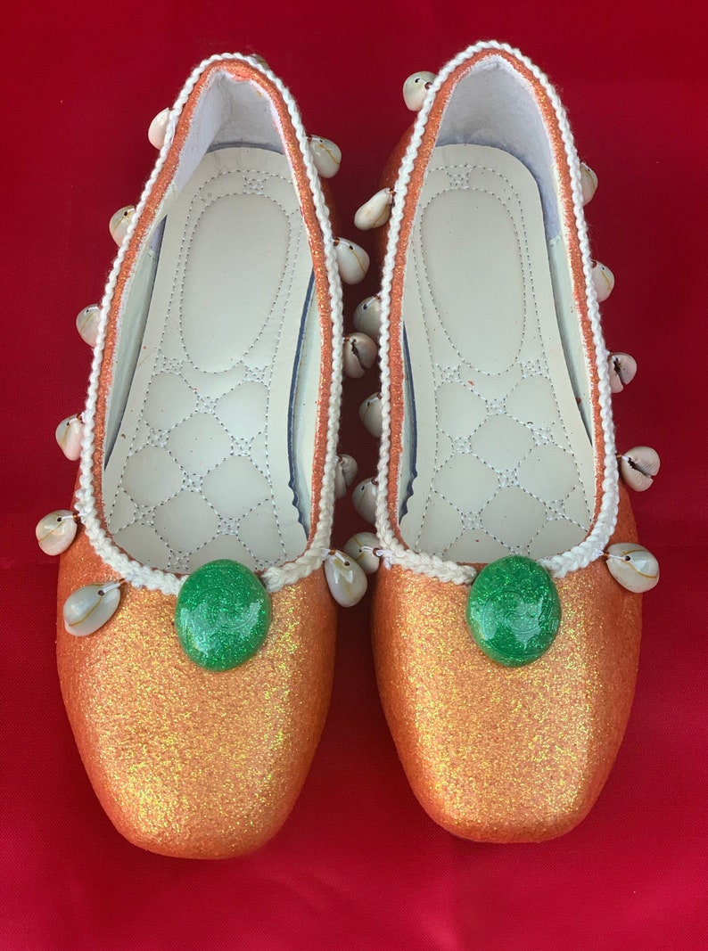 May include: A pair of orange glitter ballet flats with white trim and seashell embellishments. Each shoe has a green gem in the center.