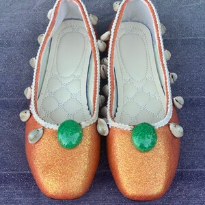 May include: A pair of orange glitter flats with white shell embellishments and green round buttons. The shoes have a white trim around the edges.