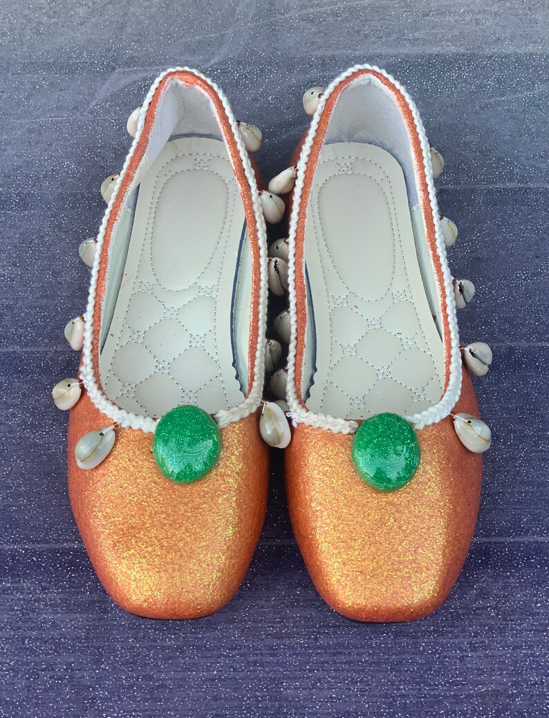 May include: A pair of orange glitter flats with white shell embellishments and green round buttons. The shoes have a white trim around the edges.