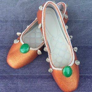 May include: A pair of orange glitter ballet flats with white trim and green stones. The flats have small white shells around the edges.