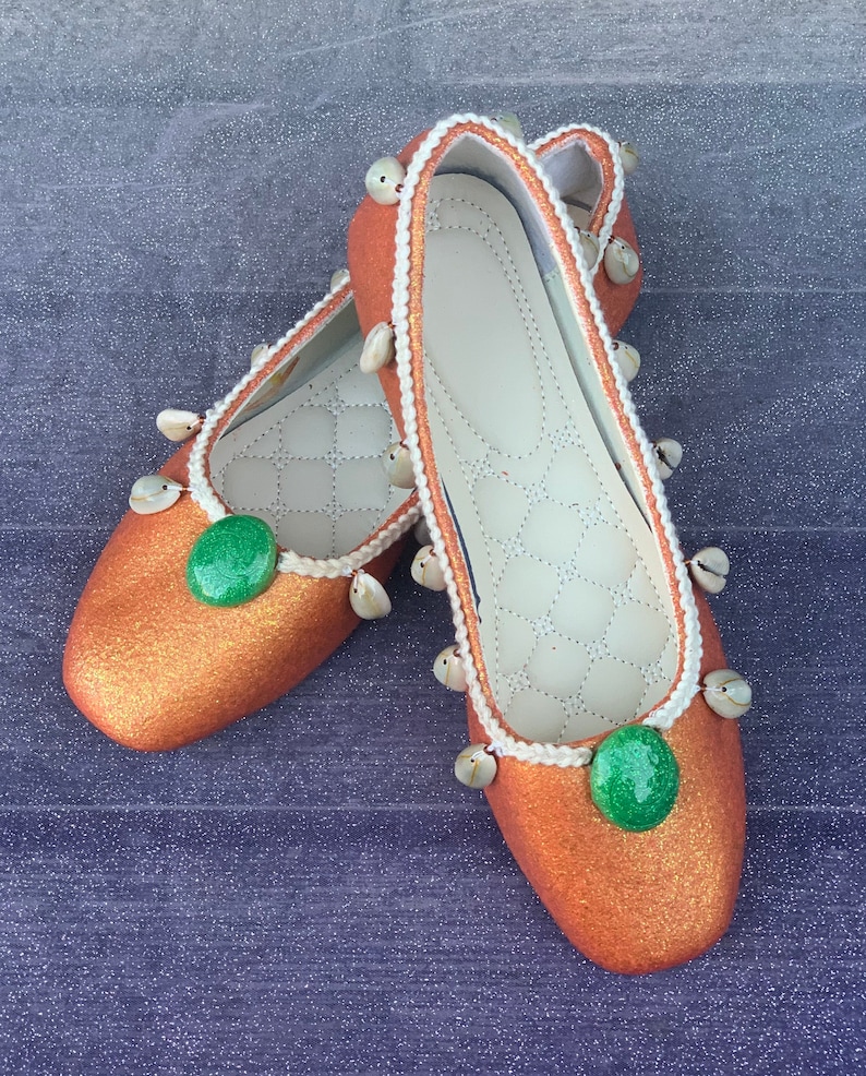 May include: A pair of orange glitter ballet flats with white trim and green stones. The flats have small white shells around the edges.