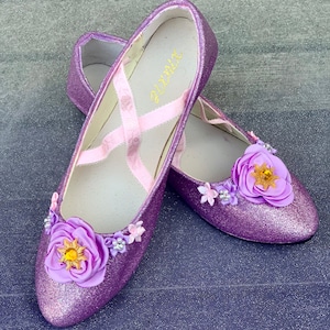 Könnte beinhalten: Ein Paar violette Glitzer-Ballerinas mit rosa Schleifenbändern und Blumenverzierungen. Die Blumen sind aus violettem Stoff mit gelber Mitte.