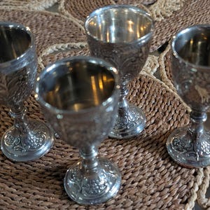 Puede incluir: Cuatro copas plateadas ornamentadas con diseños detallados de flores y volutas. Las copas tienen forma de cuenco redondeado y se asientan sobre una base de pedestal decorativa. Las copas están dispuestas sobre una superficie tejida y texturizada.