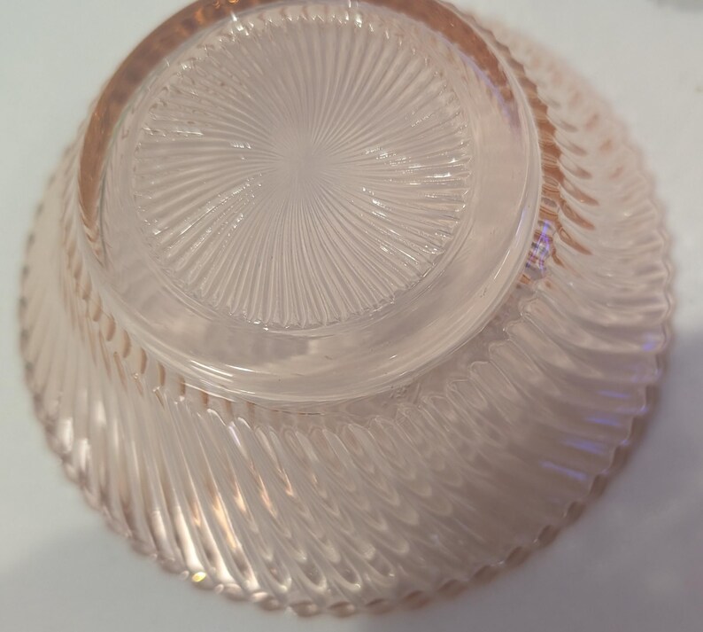 May include: A clear, pink-tinted glass bowl with a spiral design on the exterior and a starburst pattern on the base. The bowl's rim is slightly thicker than the rest of the glass, and the bowl is turned upside down.