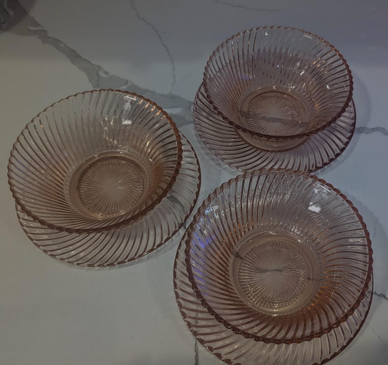 May include: Three pink glass bowls and matching plates. The bowls and plates have a spiral design. The bowls are stacked on top of the plates. The glass is a light pink color.