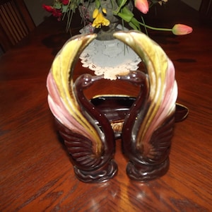 Puede incluir: Un par de saleros y pimenteros de cerámica en forma de cisnes con detalles en marrón, rosa y amarillo. Los cisnes se miran con el cuello curvado en forma de corazón.