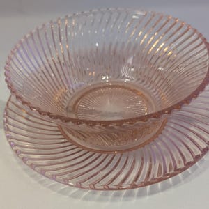 May include: A pink glass bowl and saucer set. The bowl and saucer have a ribbed design radiating from the center. The rim of the bowl has a scalloped edge. The glass has a slight pink hue.
