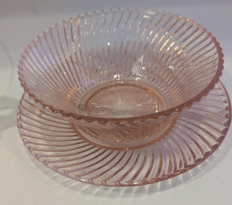 May include: A pink glass bowl and saucer set. The bowl and saucer have a ribbed design radiating from the center. The rim of the bowl has a scalloped edge. The glass has a slight pink hue.