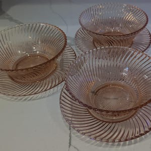 May include: Set of three pink glass bowls with matching plates. Each bowl and plate features a spiral ribbed design. The bowls are round and sit atop the plates. The glass has a translucent quality.