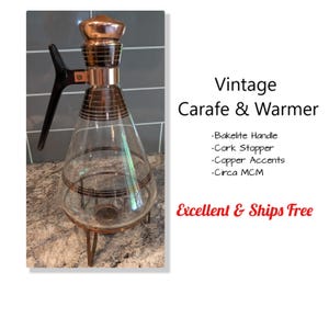 May include: Vintage glass carafe and warmer with copper accents. Features a black Bakelite handle and a copper-colored lid. The carafe has a conical shape and sits on a metal stand with three legs. The text "Vintage Carafe & Warmer" is visible.