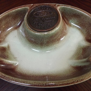 Op de afbeelding: Een groene en bruine asbak van keramiek met een verhoogd middengedeelte. Het middengedeelte heeft een metalen medaillon met de tekst "Pacific Coast Pottery, Hand Crafted, California, USA".