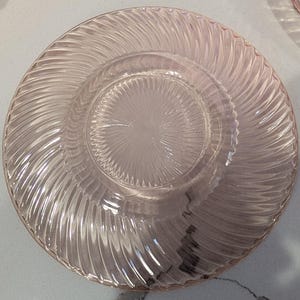 May include: A clear, pink glass plate with a spiral design around the rim and a textured center. The plate has a circular shape and is made of transparent glass. The design is visible throughout the plate.