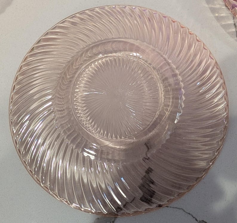 May include: A clear, pink glass plate with a spiral design around the rim and a textured center. The plate has a circular shape and is made of transparent glass. The design is visible throughout the plate.