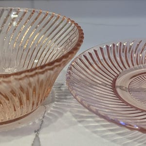 May include: A pink glass bowl and plate set. The bowl is oval-shaped with a spiral ribbed design. The plate is round with a matching spiral pattern. Both pieces are made of transparent pink glass.