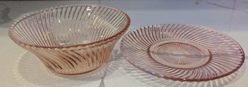 May include: A pink glass bowl and plate set. The bowl is oval-shaped with a spiral ribbed design. The plate is round with a matching spiral pattern. Both pieces are made of transparent pink glass.