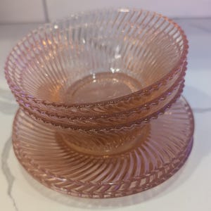 May include: A stack of three pink glass bowls and plates. The bowls and plates have a spiral ribbed design. The bowls are stacked on top of a plate. The glass is translucent, and the color is a soft pink.