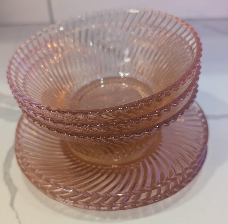 May include: A stack of three pink glass bowls and plates. The bowls and plates have a spiral ribbed design. The bowls are stacked on top of a plate. The glass is translucent, and the color is a soft pink.