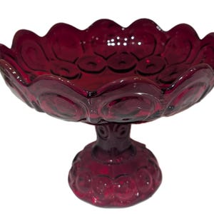 Puede incluir: Un cuenco de pedestal de vidrio rojo rubí con un borde festoneado. El cuenco tiene una base circular y un tallo decorativo. La superficie del cuenco está adornada con un patrón circular repetitivo.