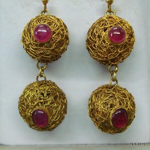 Può includere: Orecchini d'oro con due ciondoli rotondi pendenti. Ogni ciondolo è realizzato in filo d'oro intrecciato e presenta una pietra preziosa rossa al centro.