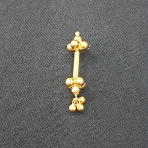 Op de afbeelding: Een goudkleurige piercing stud met een halter ontwerp. De stud heeft clusters van kleine, ronde kralen aan elk uiteinde en bungelende kralen aan de onderkant. De sieraden zijn geplaatst tegen een donkere achtergrond.