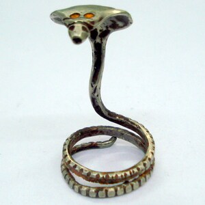 Może przedstawiać: Srebrny pierścionek w kształcie węża kobry. Głowa węża jest uniesiona, z dwoma pomarańczowymi oczami. Ciało węża owija się wokół palca, tworząc unikalną i przyciągającą wzrok biżuterię.