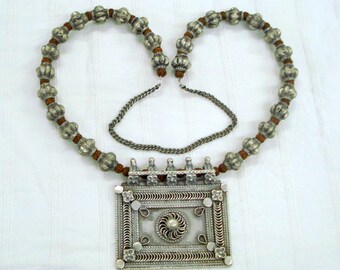 May include: A silver necklace with a large, ornate pendant. The pendant is rectangular and features intricate designs. The necklace is made of silver beads and a brown cord.