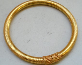 May include: A gold bangle bracelet with a textured design and a decorative band. The bracelet is made of metal and has a circular shape.