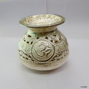 Może przedstawiać: Srebrny dzbanek z misternymi wycięciami i symbolem Om. Dzbanek ma okrągłe ciało i szeroką, płaską górę.