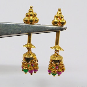 Op de afbeelding: Een paar goudkleurige oorbellen. Elke oorbel heeft een cluster van kleine gouden kralen aan de bovenkant, een decoratief gouden element en een bungelende belvormige bedel met rode en groene kralen. De oorbellen worden vastgehouden door een zilveren gereedschap.