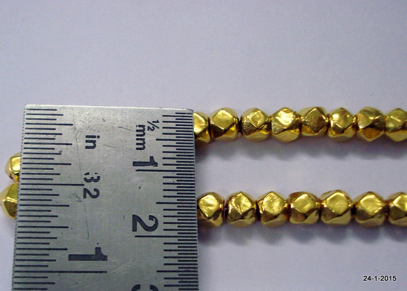 K&ouml;nnte beinhalten: Nahaufnahme einer goldfarbenen Metallperlenkette mit einem Lineal als Gr&ouml;&szlig;enreferenz. Die Perlen sind facettiert und haben eine geh&auml;mmerte Textur. Das Lineal zeigt Ma&szlig;e in Zoll und Millimetern.