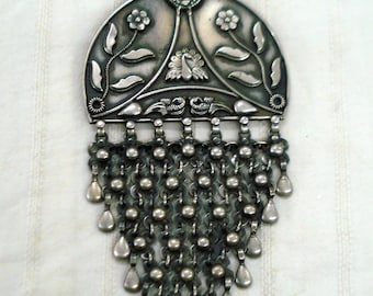 May include: A silver pendant necklace with a floral design and a dangling chain of beads. The pendant features a peacock design and is attached to a black cord.