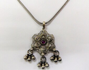 May include: Silver filigree pendant necklace with a floral design. The pendant features a central red gemstone and dangling silver beads. The necklace chain is a woven style.