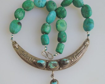 May include: A silver necklace with a crescent moon pendant and turquoise beads. The pendant has three turquoise stones and intricate filigree details.