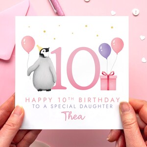 May include: A birthday card with a penguin wearing a party hat and holding a pink balloon. The card says "Happy 10th Birthday to a Special Daughter" with the name "Thea" below. Pink balloons and a gift box are also featured.