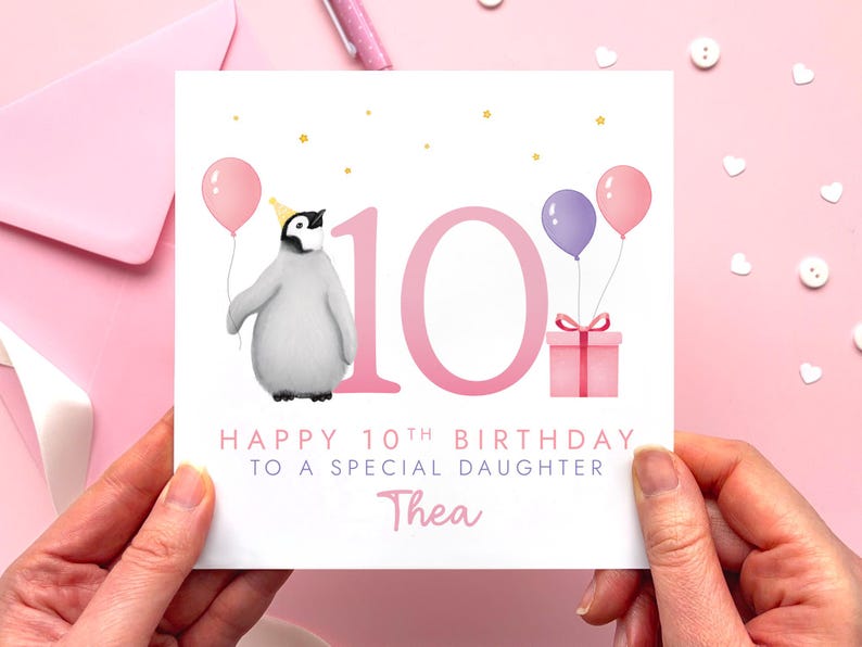 May include: A birthday card with a penguin wearing a party hat and holding a pink balloon. The card says "Happy 10th Birthday to a Special Daughter" with the name "Thea" below. Pink balloons and a gift box are also featured.