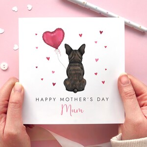 May include: A Mother's Day card featuring a seated brindle dog holding a pink heart-shaped balloon. The card reads "Happy Mother's Day Mum" with small pink hearts scattered around the dog. The card is held against a pink background.