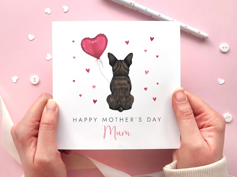 May include: A Mother's Day card featuring a seated brindle dog holding a pink heart-shaped balloon. The card reads "Happy Mother's Day Mum" with small pink hearts scattered around the dog. The card is held against a pink background.