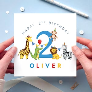 May include: A square white birthday card featuring a large blue number 2, a giraffe, a crocodile, a monkey, a lion, and a zebra wearing party hats. The text reads HAPPY 2ND BIRTHDAY and OLIVER in colourful letters with blue bunting and a red balloon.
