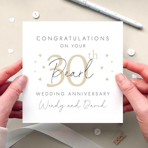 K&ouml;nnte beinhalten: Eine quadratische wei&szlig;e Gru&szlig;karte mit dem Text CONGRATULATIONS ON YOUR 30th Pearl WEDDING ANNIVERSARY Wendy and David. Das Design enth&auml;lt eine gro&szlig;e beige Zahl 30, schwarze Schreibschrift und kleine beige Herzsymbole um den zentralen Text herum.