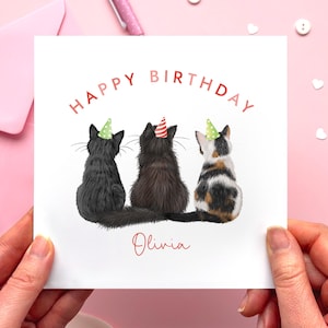 May include: A white greeting card with the words "HAPPY BIRTHDAY" in red. The card features three cats wearing party hats. The name "Olivia" is written in red script below the cats. The card is held by two hands.