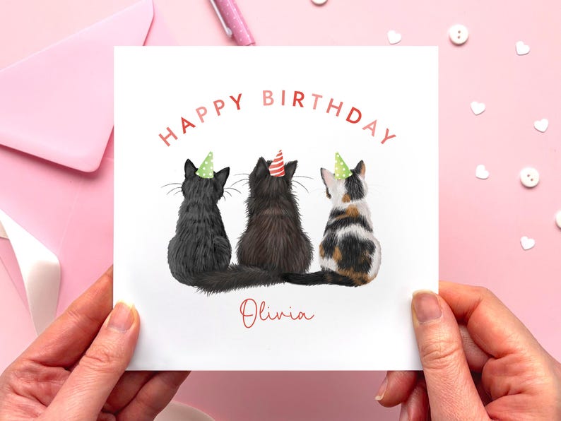 May include: A white greeting card with the words "HAPPY BIRTHDAY" in red. The card features three cats wearing party hats. The name "Olivia" is written in red script below the cats. The card is held by two hands.